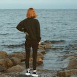 A woman stands facing the sea, hand on hip, on a rocky seashore.