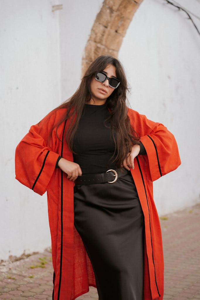 A stylish woman posing outdoors in a black dress and red coat, exuding sophistication.