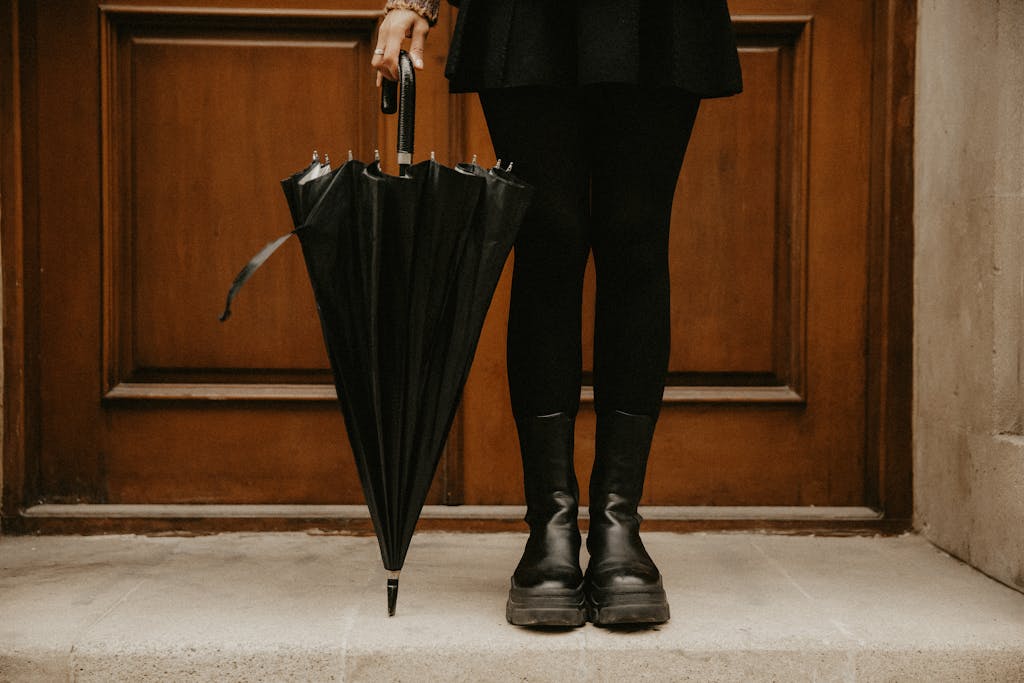 A woman holds a black umbrella, standing in front of a wooden door, showcasing chic fashion.