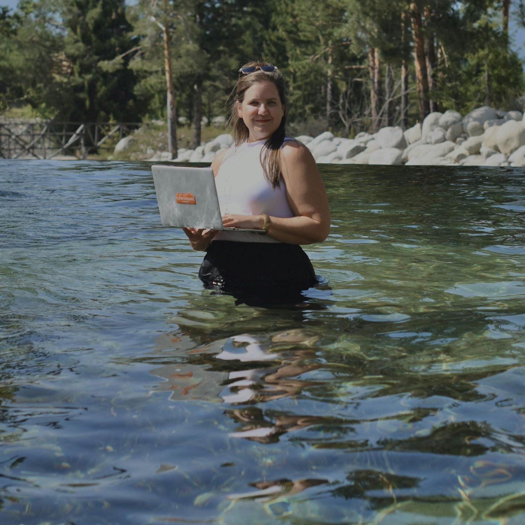 A woman works on her laptop while standing in a serene outdoor lake setting.