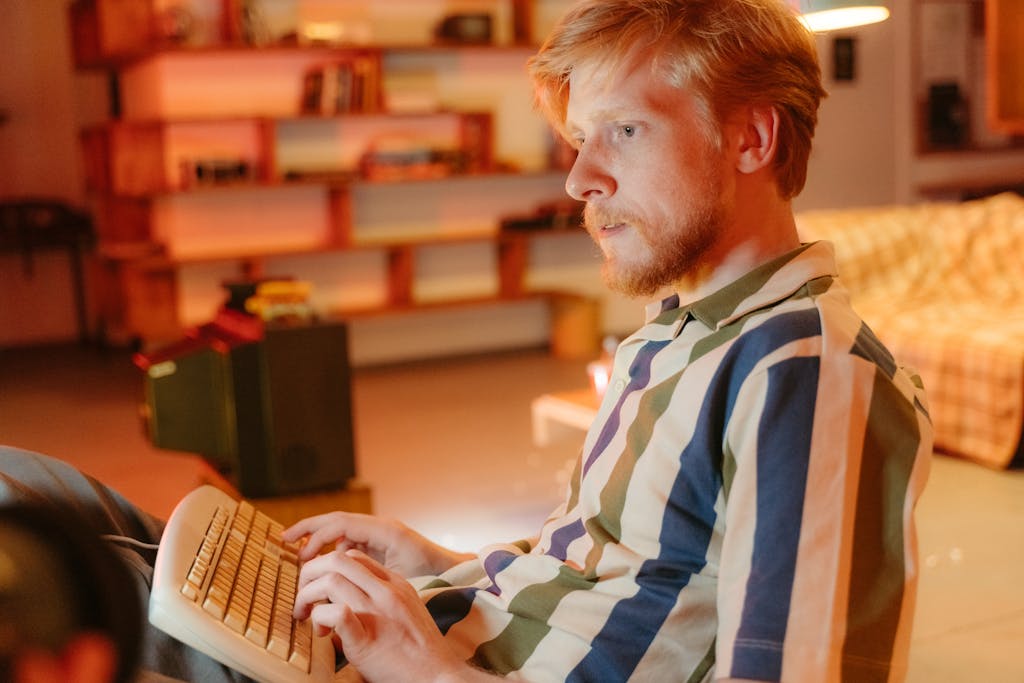 Bearded man in striped shirt typing on a vintage keyboard in a warm, indoor setting.