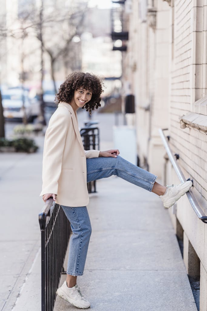 Charming young woman with curly hair posing in a trendy outfit on a city street, exuding a joyful vibe.