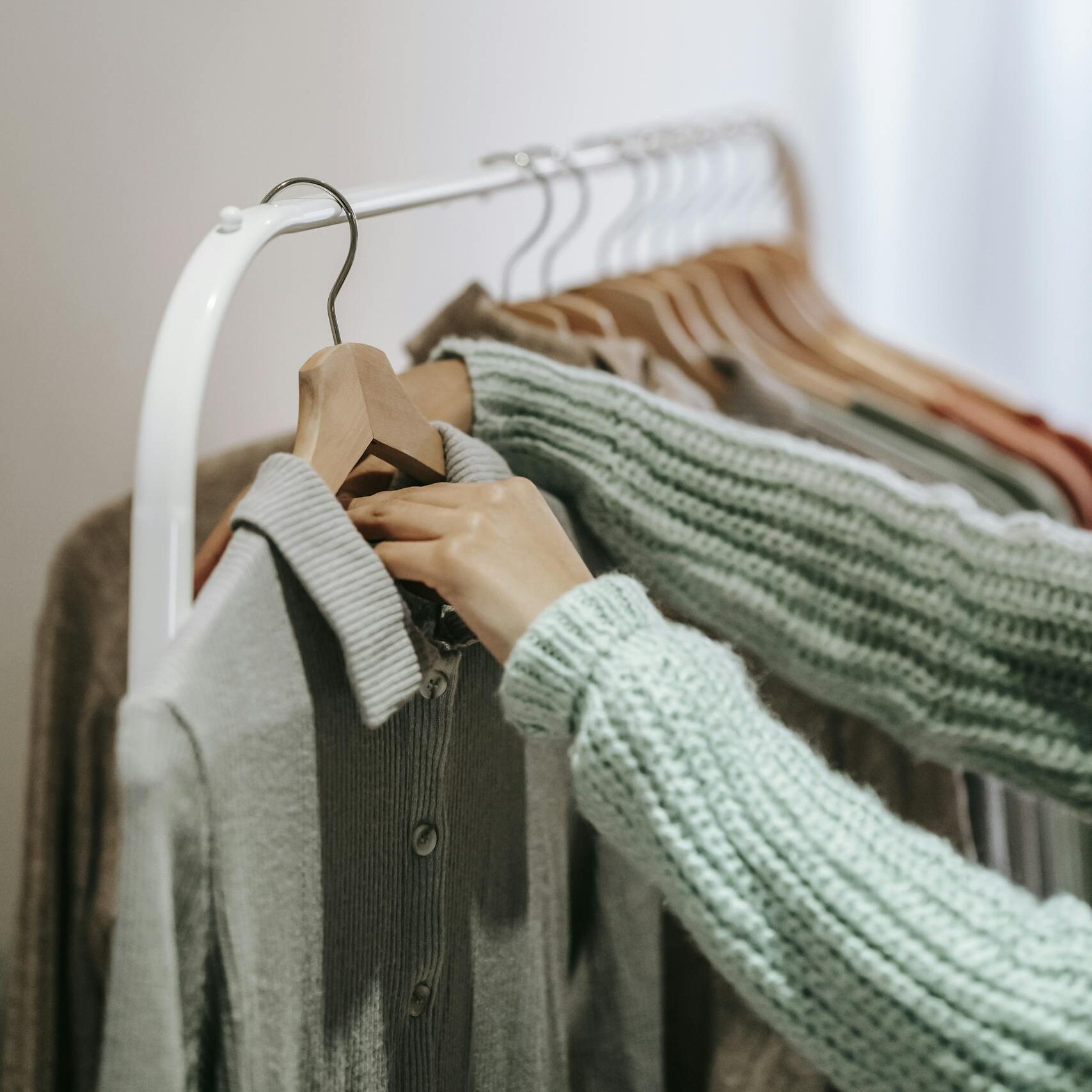 Crop unrecognizable female in knitted wear touching wooden hanger with clothes on stand in building