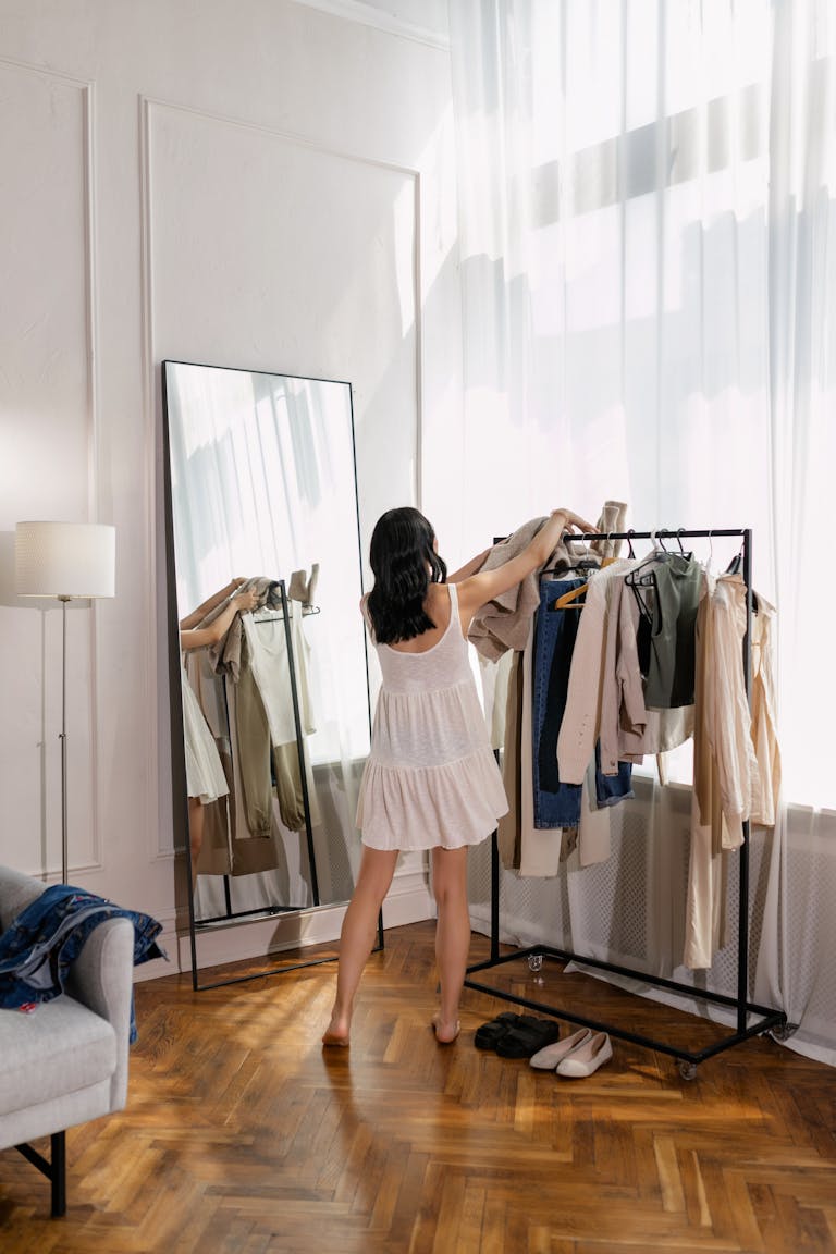 Woman in white dress organizing clothes on a clothing rack in a sunny room with wooden flooring.
