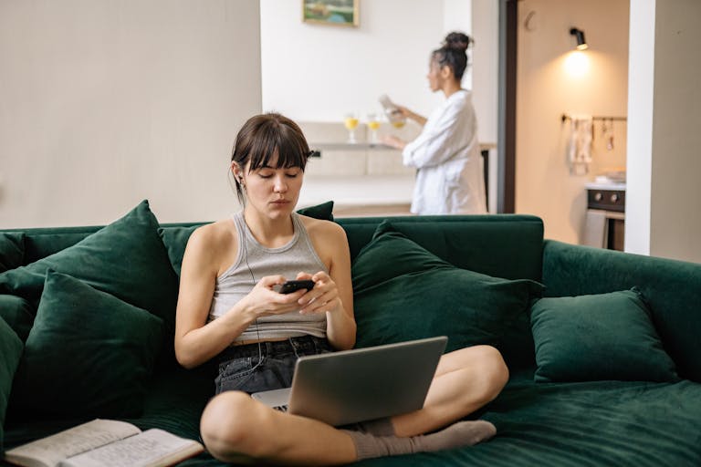 Woman sitting on a green couch using her phone and laptop while another woman prepares a drink in the background.