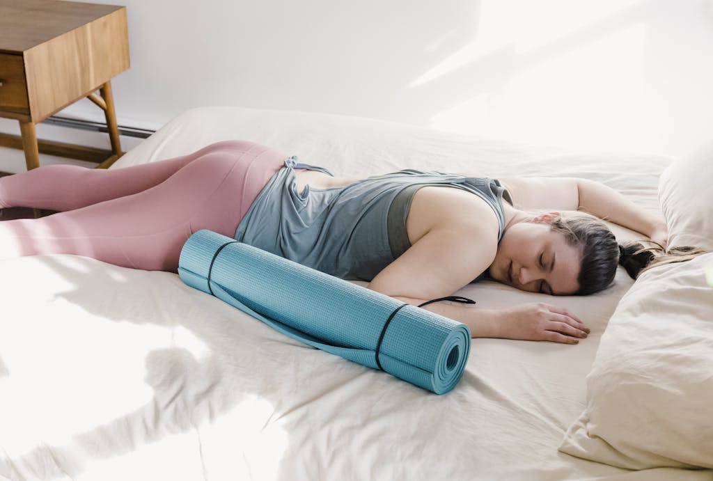 Young woman in active wear naps on bed with yoga mat, embracing relaxation in a sunlit room.
