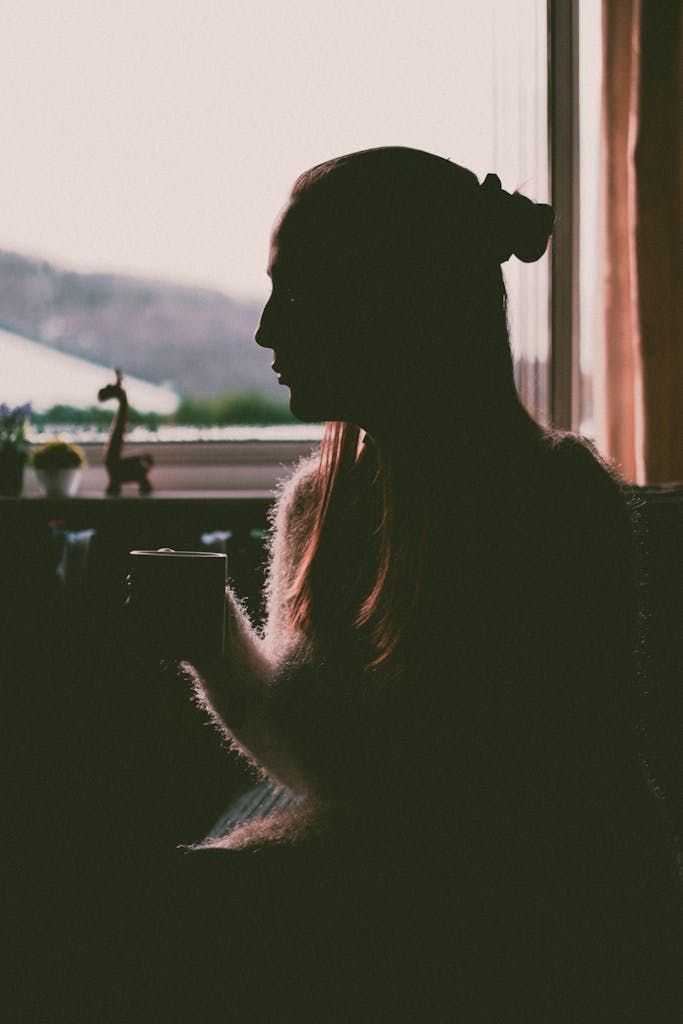 A contemplative silhouette of a woman holding a cup by the window at twilight.