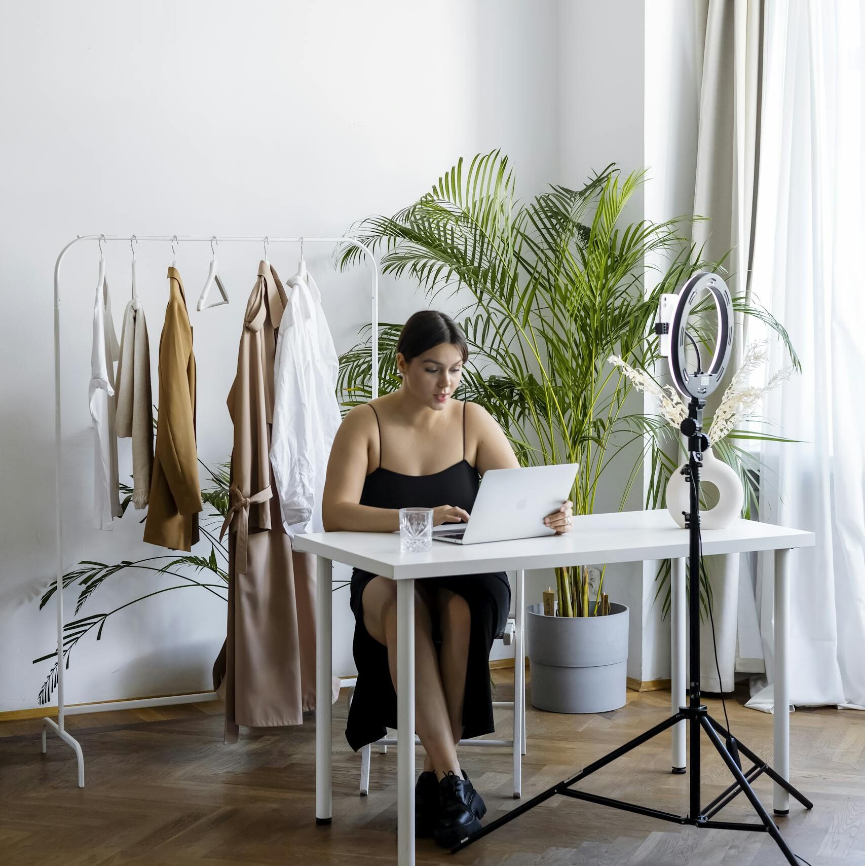 A woman engaging in online selling with clothes rack, laptop, and ring light setup indoors.