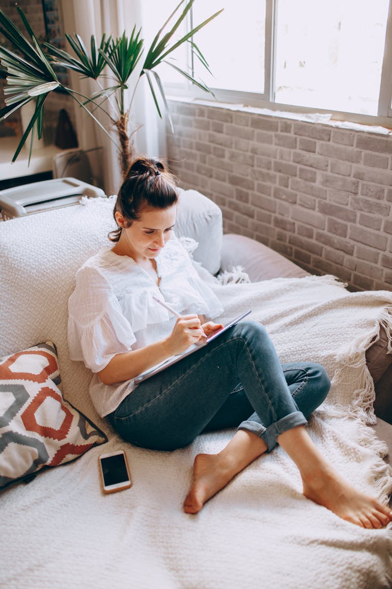 A woman sitting barefoot on a sofa writing in a notebook in a cozy, sunlit living room.
