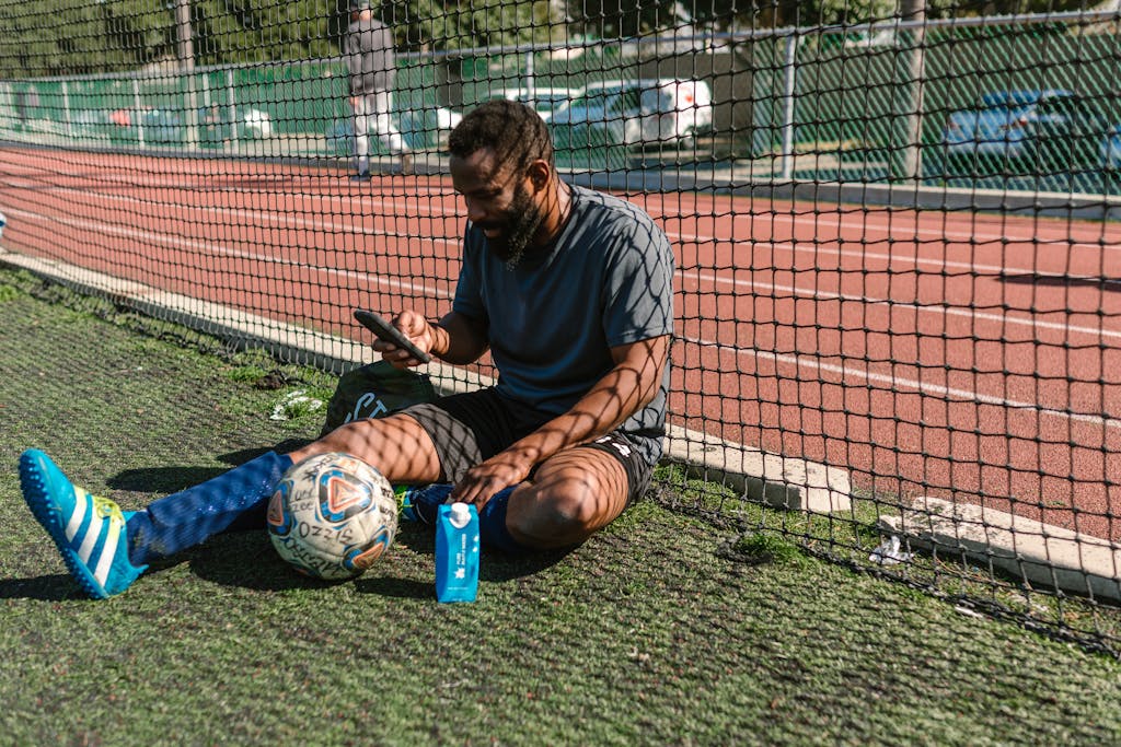 Athlete taking a break on a soccer field with ball and smartphone, enjoying leisure time.