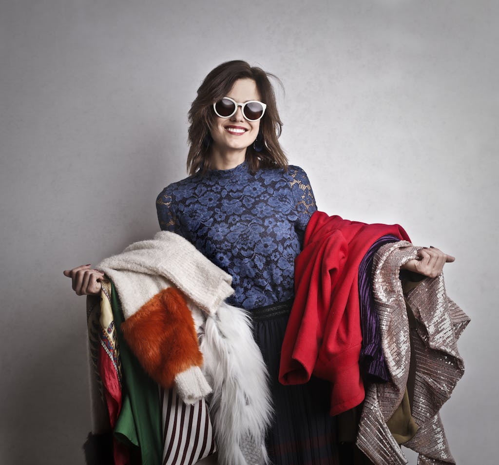 Elegant woman in sunglasses holding a variety of colorful outerwear, smiling at the camera.