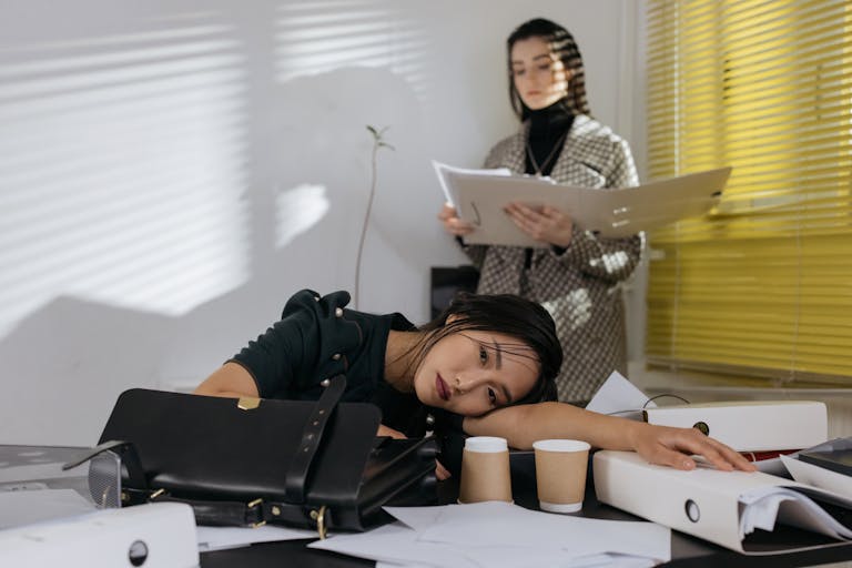 Exhausted employee resting on desk surrounded by documents, colleague in background.