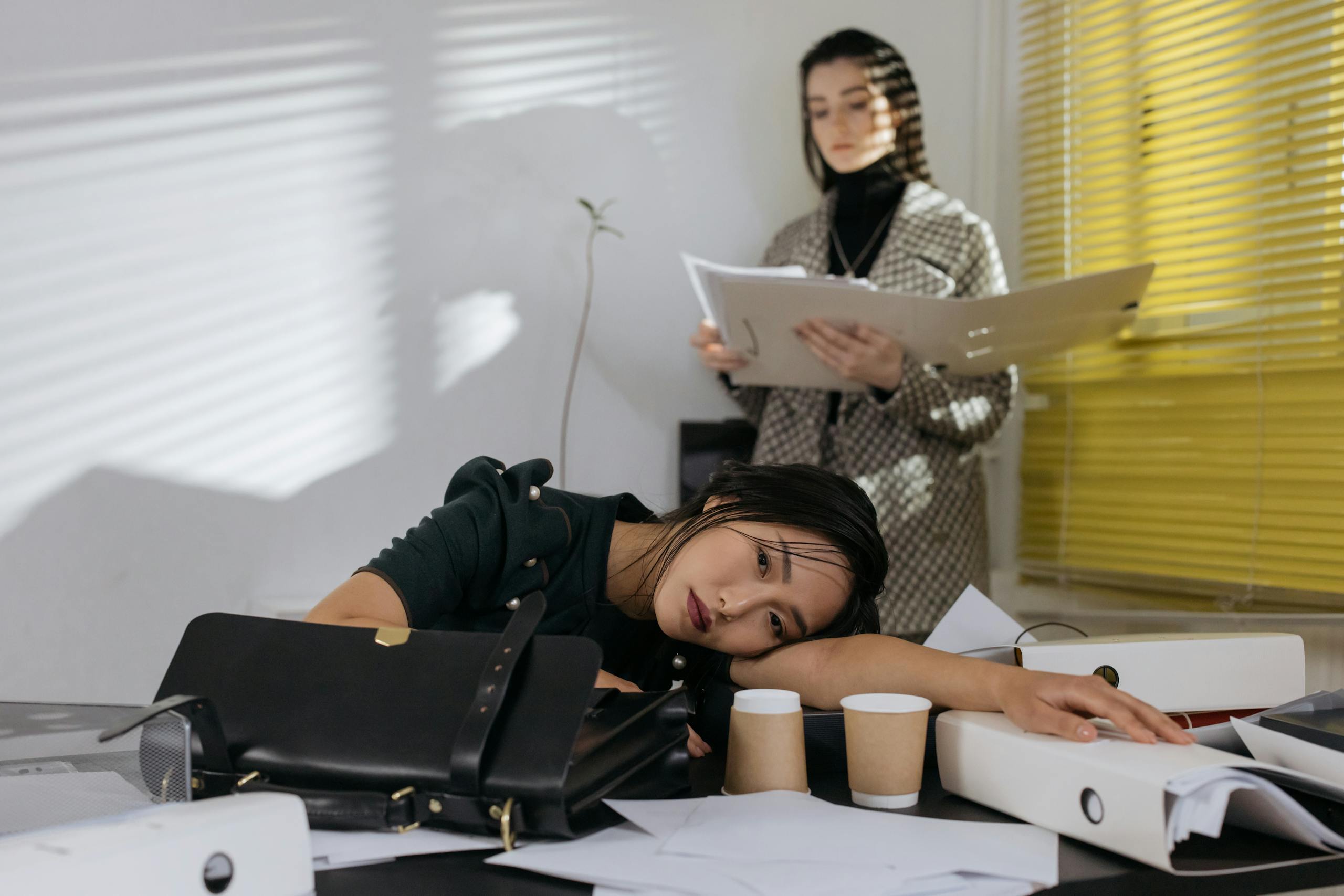 Exhausted employee resting on desk surrounded by documents, colleague in background.