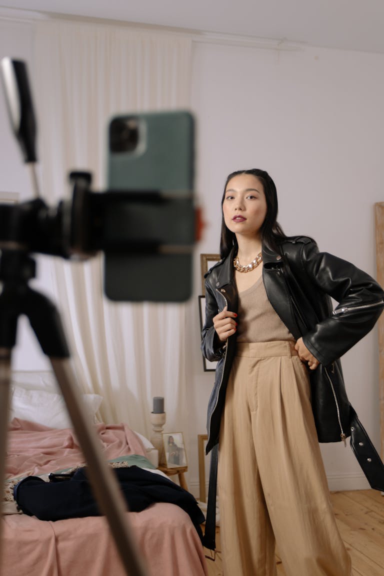 Fashionable woman posing in black leather jacket indoors, capturing content with smartphone and tripod.