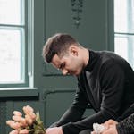 A young man in black holds flowers, symbolizing loss and mourning, seated indoors.